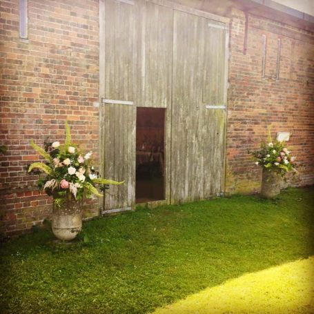 Rustic barn entrance with a small doorway, flanked by floral arrangements in pots.